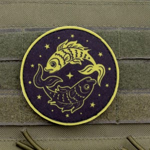 May include: A round patch featuring a yellow and navy blue Pisces zodiac design. Two stylized fish are depicted, facing opposite directions, surrounded by stars. The patch has a yellow border and is set against a green background.