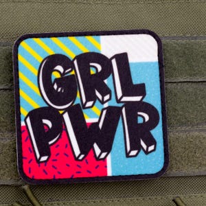May include: A square patch with the text "GRL PWR" in bold, white, 3D letters. The background is divided into four sections: yellow and blue stripes, white, blue, and red with pink dots. The patch is on a green fabric.