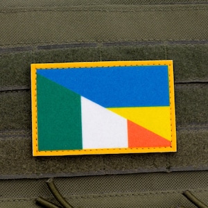Puede incluir: Un parche rectangular con borde amarillo, que presenta un diseño de bandera con secciones verdes, blancas, azules, amarillas y naranjas. El parche está adherido a una superficie de tela verde, probablemente un chaleco táctico o equipo similar.