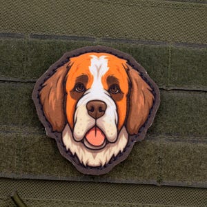 Puede incluir: Un parche colorido con un perro San Bernardo de dibujos animados con pelaje marrón, blanco y naranja. El perro tiene una expresión amigable con la lengua fuera. El parche está adherido a un fondo de tela verde.