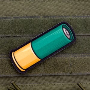 Op de afbeelding: Een patch met een shotgun cartridge, met een groene behuizing en een gele basis, omlijnd in zwart. De patch is geplaatst op een groene, getextureerde ondergrond, mogelijk een tactisch vest of uitrusting.