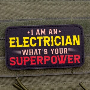 May include: A rectangular patch with the text "I AM AN ELECTRICIAN WHAT'S YOUR SUPERPOWER" in yellow and red lettering on a black background. The patch is attached to a green fabric.