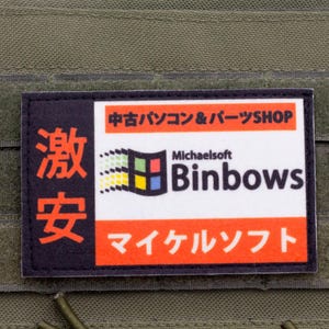 Puede incluir: Un parche rectangular con un lado izquierdo negro con texto japonés en rojo y un lado derecho blanco con barras naranjas. El centro muestra un logotipo de ventana multicolor, el texto Michaelsoft Binbows y caracteres japoneses.