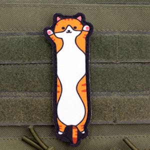 May include: A rectangular patch featuring a cartoon cat with orange and white fur, arms raised. The patch has a black border and is affixed to a green fabric background with hook and loop fasteners.