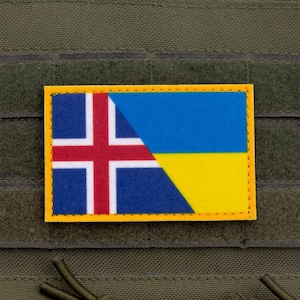 Può includere: Una toppa rettangolare con le bandiere di Islanda e Ucraina. La bandiera islandese, a sinistra, ha uno sfondo blu con una croce rossa bordata di bianco. La bandiera ucraina, a destra, ha strisce blu e gialle. La toppa ha un bordo giallo.