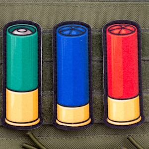 以下が含まれることがあります： カラフルな散弾銃シェルパッチ3枚。パッチは緑、青、赤で、金色のベースと黒い縁取りがあります。パッチは緑色のタクティカルベストに表示されています。
