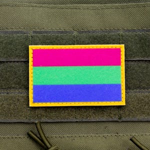 May include: A rectangular patch with a yellow border, featuring horizontal stripes of pink, green, and blue. The patch is affixed to a dark green fabric with a hook and loop fastener.
