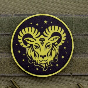 Può includere: Una toppa circolare con il disegno di una testa di ariete gialla con corna ricurve su sfondo viola scuro. Il design include piccole stelle gialle sparse attorno all'ariete. La toppa ha un bordo giallo ed è posta su un tessuto verde oliva.
