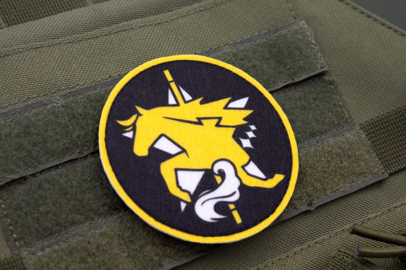 Puede incluir: Un parche bordado amarillo y blanco con un dise&ntilde;o de caballo estilizado sobre un fondo negro. El parche est&aacute; sujeto a una superficie de tela verde.