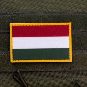May include: A rectangular patch displaying the Hungarian flag, with horizontal stripes of red, white, and green, set within a yellow border. The patch is attached to a textured, olive-green fabric.