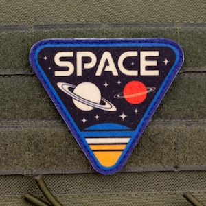 May include: A triangular patch with the word "SPACE" in white letters, featuring a planet with rings and a red planet. The patch has a blue border and is set against a green fabric background.