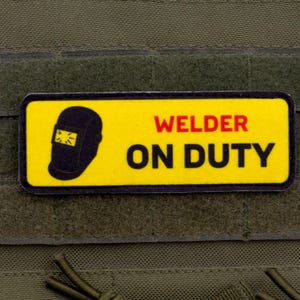 Könnte beinhalten: Ein rechteckiger Aufnäher mit den Worten "WELDER ON DUTY" in roter und schwarzer Schrift auf gelbem Hintergrund. Links ist eine schwarze Schweißmaske abgebildet. Der Aufnäher ist auf einem grünen Stoff befestigt.