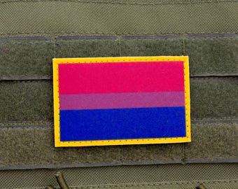 May include: A rectangular patch featuring the bisexual pride flag, with horizontal stripes in pink, purple, and blue, outlined in yellow. The patch is affixed to a green, textured fabric background.