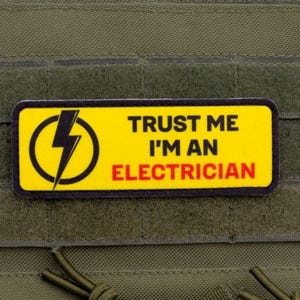 Puede incluir: Un parche rectangular con fondo amarillo y borde negro. El parche presenta un rayo negro dentro de un círculo amarillo a la izquierda. El texto "TRUST ME I'M AN ELECTRICIAN" está impreso en negro y rojo.