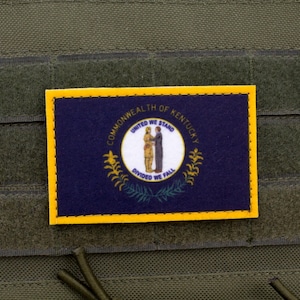 May include: A rectangular patch featuring the Kentucky state flag, with a navy blue background and a gold border. The flag displays the state seal with the text "COMMONWEALTH OF KENTUCKY" and "UNITED WE STAND, DIVIDED WE FALL". The patch is affixed to a green fabric.
