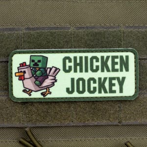 May include: A rectangular patch with the words "CHICKEN JOCKEY" in dark green block letters. The patch features a cartoon image of a pixelated green figure riding a chicken. The patch is on a green fabric background.