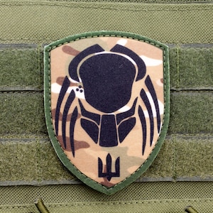 May include: A green camouflage patch featuring a black silhouette of a predator alien head and the Ukrainian trident symbol.