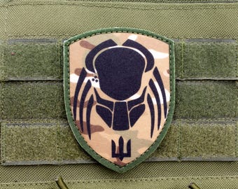 Op de afbeelding: Een groene camouflage patch met een zwart silhouet van een Predator alien hoofd en het Oekraïense drietand symbool.