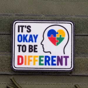 Könnte beinhalten: Ein rechteckiger Aufnäher mit weißem Hintergrund und schwarzem Rand. Er zeigt den Text IT'S OKAY TO BE DIFFERENT in Blau, Schwarz und Regenbogenfarben. Rechts ist der schwarze Umriss eines menschlichen Kopfes mit einem Herz aus Puzzleteilen.
