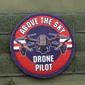May include: A round patch with the text "ABOVE THE SKY" and "DRONE PILOT" encircling a drone illustration. The patch has a red and blue border and is set against a green fabric background.