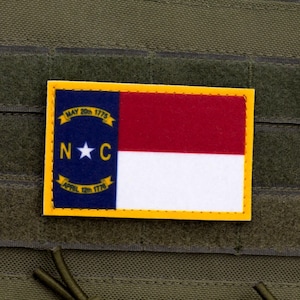 May include: A yellow-bordered patch with the North Carolina state flag. The flag features a red and white stripe with a blue triangle in the upper left corner. The triangle has a white star and the letters "N.C." in white. The text "MAY 20th 1775" and "APRIL 12th 1776" are on the blue triangle.
