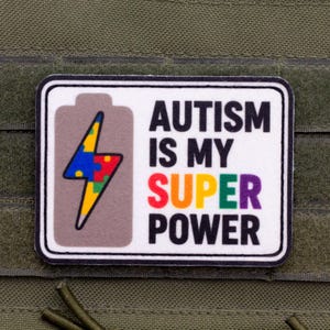 Puede incluir: Un parche rectangular con fondo blanco y borde negro. Presenta un icono de batería gris que contiene un rayo relleno con un patrón de piezas de rompecabezas multicolor. El texto dice AUTISM IS MY SUPER POWER en colores negro y arcoíris.
