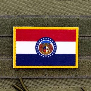 以下が含まれることがあります： ミズーリ州旗をあしらった長方形のパッチ。赤、白、青のデザインで、中央に円形のデザインのエンブレムがあり、黄色のフレームで縁取られています。パッチは緑色の生地に取り付けられています。