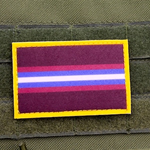 May include: A rectangular patch with a yellow border, featuring a dark burgundy background with horizontal stripes of red, white, and blue. The patch is affixed to a green fabric with hook-and-loop fasteners, commonly used on tactical gear.