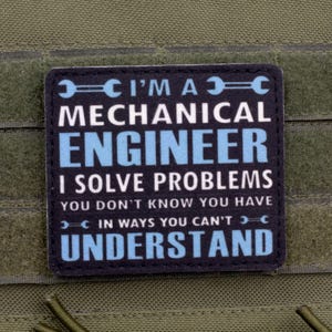Op de afbeelding: Een zwart en blauw embleem met de tekst "I'M A MECHANICAL ENGINEER I SOLVE PROBLEMS YOU DON'T KNOW YOU HAVE IN WAYS YOU CAN'T UNDERSTAND". Het embleem is rechthoekig en bevestigd aan een groene stof.