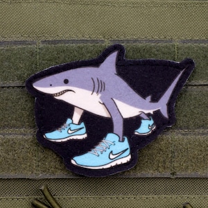 Può includere: Una toppa con un cartone animato di uno squalo che indossa scarpe da ginnastica azzurre con suole bianche e un logo Nike bianco. Lo squalo è grigio e bianco, su sfondo nero, ed è fissato a un tessuto verde.