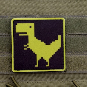 Pode incluir: Um patch quadrado com um dinossauro amarelo pixelizado sobre um fundo roxo escuro, emoldurado por uma borda amarela. O patch é preso a uma superfície de tecido verde com velcro.