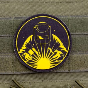 Può includere: Toppa rotonda con un disegno giallo e nero di un saldatore con maschera e scintille. La toppa è su uno sfondo verde con velcro, progettata per essere attaccata a equipaggiamento tattico o abbigliamento.