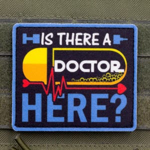 May include: A square morale patch with a black background and a blue border. The text reads IS THERE A DOCTOR HERE? in white and blue. A yellow and white pill graphic is centred with a red EKG heart rate line running through the middle.