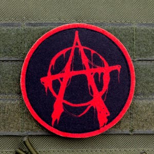 Op de afbeelding: Een ronde patch met een rood anarchiesymbool op een zwarte achtergrond, met een rode rand. De patch is bevestigd op een groene stof met klittenband, vaak gebruikt op tactische uitrusting.