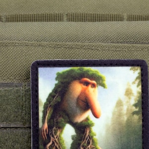 May include: A rectangular patch featuring a whimsical illustration of a creature with a long nose and leafy hair, set against a forest backdrop. The patch is bordered in black and is affixed to a green fabric.