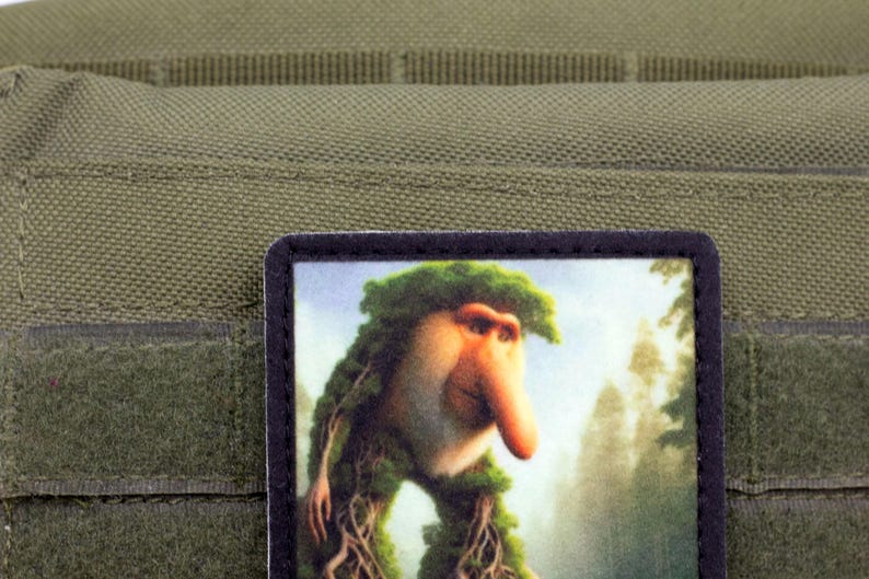 May include: A rectangular patch featuring a whimsical illustration of a creature with a long nose and leafy hair, set against a forest backdrop. The patch is bordered in black and is affixed to a green fabric.