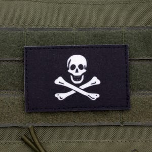 Może przedstawiać: Czarna łatka z białym wzorem czaszki i skrzyżowanych kości. Łatka jest przymocowana do zielonego taktycznego kamizelki.