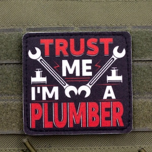 Puede incluir: Un parche negro y rojo con el texto "TRUST ME I'M A PLUMBER" en blanco. El parche presenta dos llaves inglesas blancas y un diseño de rayo. El parche está sobre un fondo de tela verde.