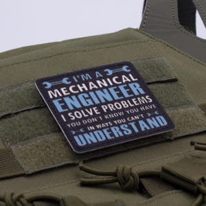 May include: A square patch with the text "I'M A MECHANICAL ENGINEER I SOLVE PROBLEMS YOU DON'T KNOW YOU HAVE IN WAYS YOU CAN'T UNDERSTAND" in white and blue on a black background. The patch is affixed to an olive green tactical vest.