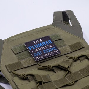 K&ouml;nnte beinhalten: Olivgr&uuml;ne taktische Weste mit einem Aufn&auml;her mit der Aufschrift "I'M A PLUMBER TO SAVE TIME JUST ASSUME I'M ALWAYS RIGHT" in wei&szlig;er und blauer Schrift. Die Weste hat Riemen und Schlaufen.