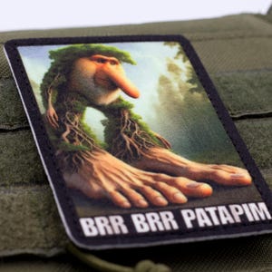 May include: A rectangular patch featuring a whimsical creature with a long nose, leafy hair, and large hands. The patch has the text "BRR BRR PATAPIM" in white letters on a black background. The patch is set against a green background.
