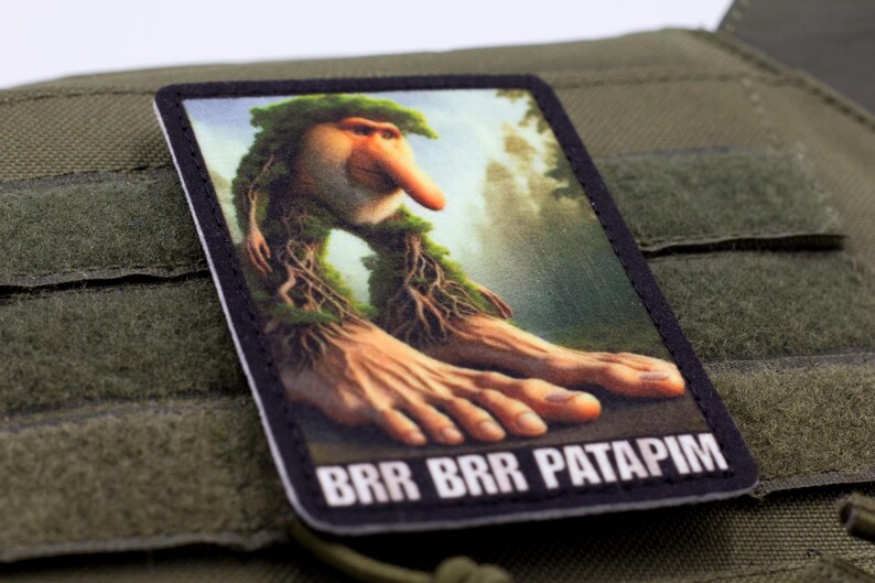 May include: A rectangular patch featuring a whimsical creature with a long nose, leafy hair, and large hands. The patch has the text "BRR BRR PATAPIM" in white letters on a black background. The patch is set against a green background.