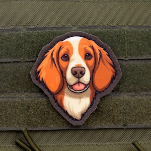May include: A patch featuring a cartoon beagle dog with orange and white fur, a black outline, and a pink tongue. The patch is attached to a green fabric background with hook and loop fasteners.