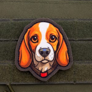 Op de afbeelding: Een kleurrijke patch met het gezicht van een beagle-hond. De hond heeft oranje en witte vacht, met grote bruine ogen en een blauwe neus. De patch is geplaatst tegen een groene achtergrond met klittenband.