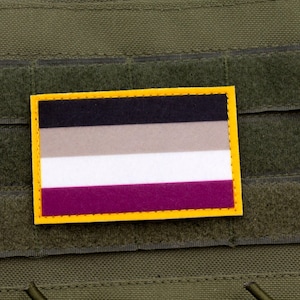 Può includere: Una toppa rettangolare con bordo giallo, raffigurante una bandiera a strisce orizzontali nere, grigie, bianche e viola. La toppa è fissata su una superficie di tessuto verde con chiusure a strappo.