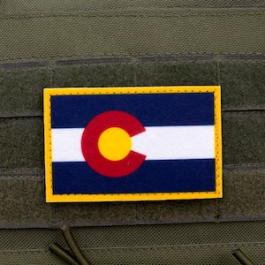 May include: A rectangular patch displaying the Colorado state flag. The flag features a blue background, a white stripe, a red 'C' with a gold disc, and a yellow border. The patch is attached to a green fabric.