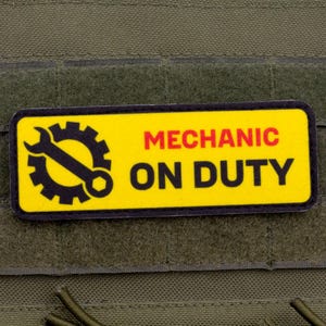 Può includere: Una toppa gialla e nera con le parole "MECHANIC ON DUTY" in rosso e nero. La toppa presenta un'icona nera di ingranaggio e chiave inglese. La toppa è attaccata a un tessuto verde.