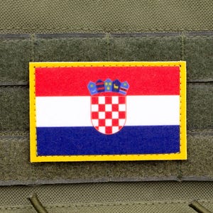 Può includere: Una toppa rettangolare con la bandiera croata, con un tricolore orizzontale rosso, bianco e blu e lo stemma croato. La toppa è bordata di giallo e attaccata a un tessuto verde.