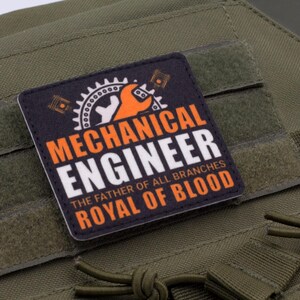 Pu&ograve; includere: Una toppa quadrata con le parole "MECHANICAL ENGINEER" in bianco e arancione, insieme al testo "THE FATHER OF ALL BRANCHES ROYAL OF BLOOD" su sfondo nero. La toppa &egrave; posta su un tessuto verde.