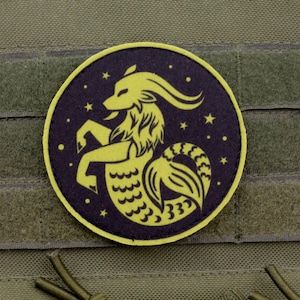 Op de afbeelding: Een ronde patch met het sterrenbeeld Steenbok, een geiten-vis hybride, in geel tegen een donkerblauwe achtergrond met sterren. De patch is bevestigd op een groene stoffen ondergrond met klittenbandsluitingen.